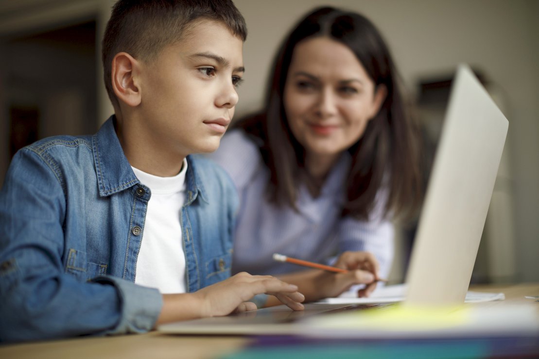 Schüler konzentriert am Laptop, Mutter im Hintergrund